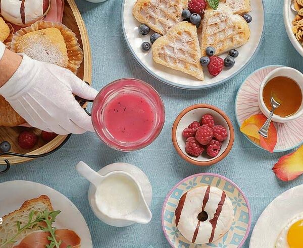 A chef serving a Mother's Day brunch spread with waffles, berries, croissants, eggs, and a pink drink on a light blue table