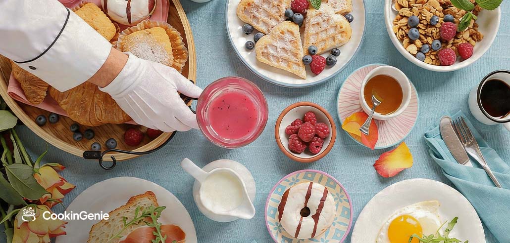 A chef serving a Mother's Day brunch spread with waffles, berries, croissants, eggs, and a pink drink on a light blue table